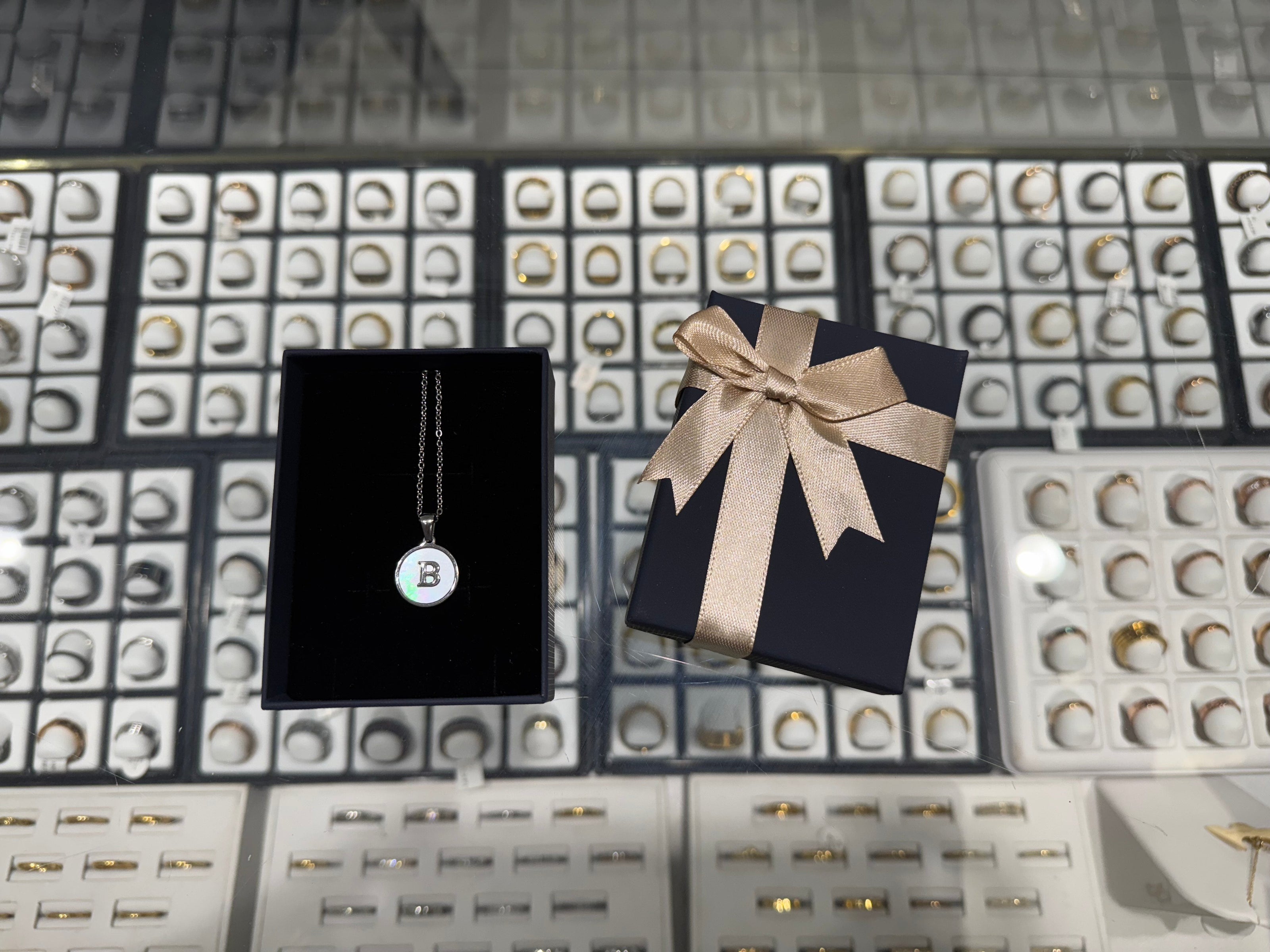 Necklace with a 'B' pendant on a black card, next to a gift box with a bow, against a jewelry display background.