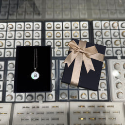 Necklace with a 'B' pendant on a black card, next to a gift box with a bow, against a jewelry display background.