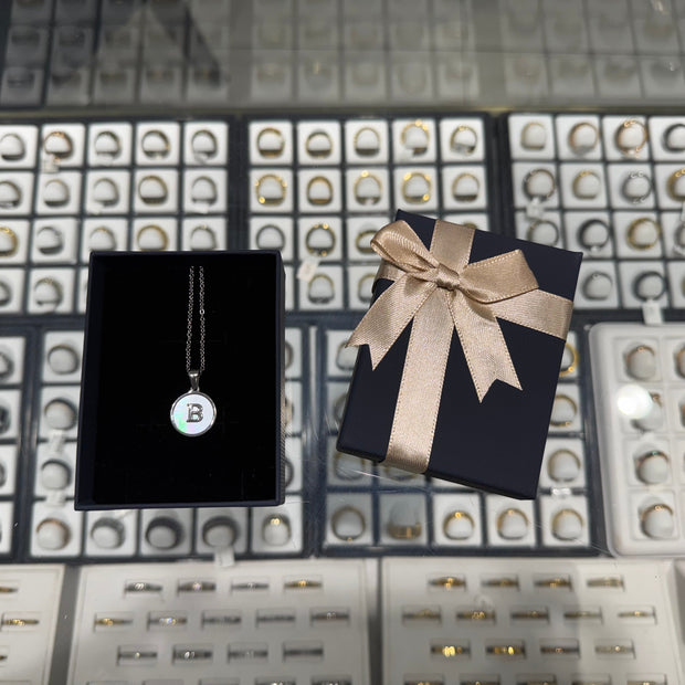Necklace with a 'B' pendant on a black card, next to a gift box with a bow, against a jewelry display background.